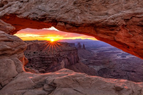 Sunset view through a rock arch, illuminating rugged cliffs and valleys in a desert landscape.