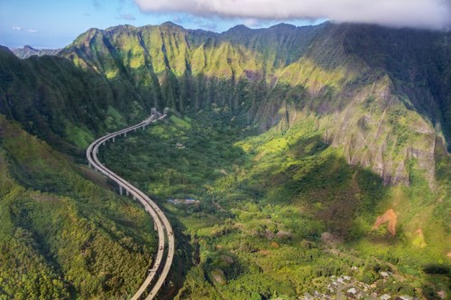 Aerial view of a winding highway through lush green mountains under a partly cloudy sky.