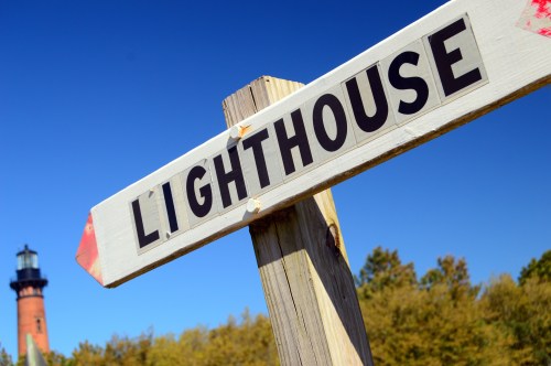 A wooden sign pointing to a lighthouse, with the lighthouse visible in the background against a clear blue sky.
