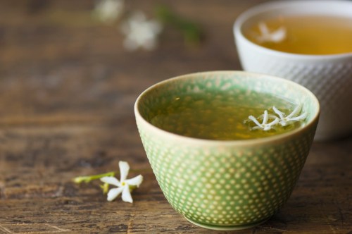 A green bowl filled with tea, garnished with white flowers, sits on a wooden table next to a lighter-colored cup.