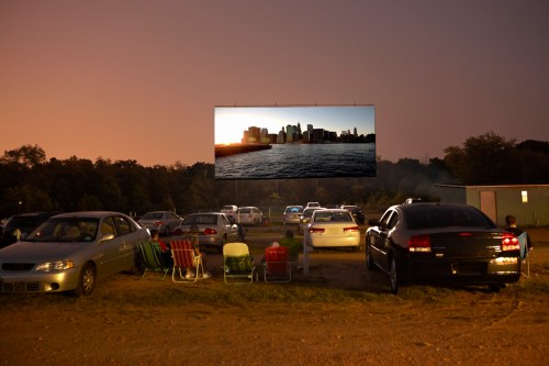 Cars and lawn chairs are set up in a field, facing a large outdoor screen showing a city skyline at sunset.