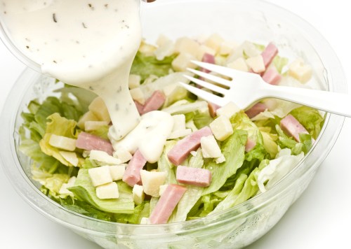 A hand pours dressing over a salad with lettuce, cheese cubes, and ham, served in a clear bowl with a fork.