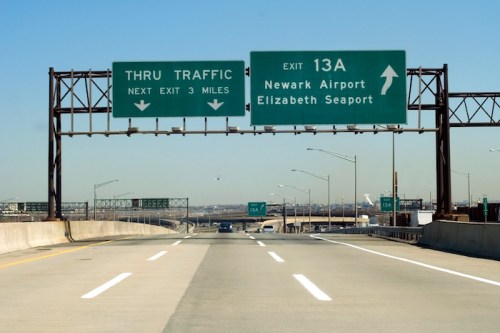 Highway sign indicating "Thru Traffic" and exit 13A for Newark Airport and Elizabeth Seaport, with clear blue sky.