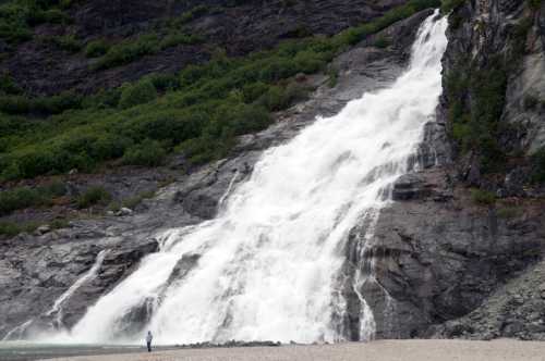 A person stands near a large waterfall cascading down rocky cliffs, surrounded by lush greenery.