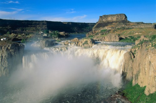 A stunning waterfall cascades over rocky cliffs, surrounded by lush greenery and a clear blue sky.