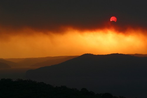 A dramatic sunset over mountains, with the sun partially obscured by dark clouds, creating a fiery orange glow.