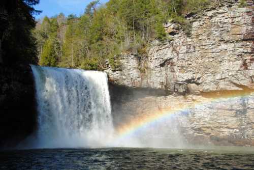 A waterfall cascades over rocky cliffs, creating mist and a vibrant rainbow in the sunlight.