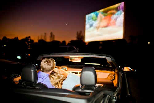A couple cuddles in a convertible at a drive-in movie, watching a film on a large screen as the sun sets.