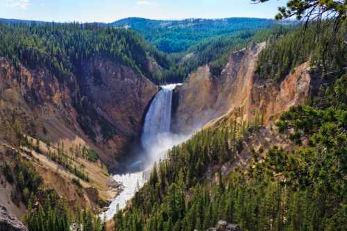 A stunning waterfall cascades down a rocky cliff, surrounded by lush green forests and mountains under a clear blue sky.