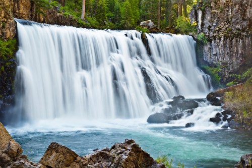 A serene waterfall cascades over rocks into a clear blue pool, surrounded by lush green trees and natural scenery.