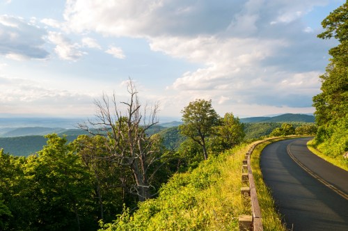 A winding road curves through lush green hills under a partly cloudy sky, with distant mountains visible in the background.