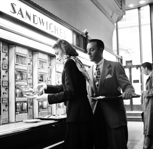A woman selects food from a vintage sandwich vending machine while a man holds a tray behind her in a retro café setting.