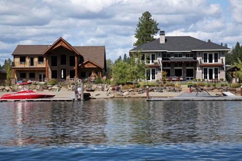 Two large houses by a lake, one with a wooden facade and the other modern, with a boat docked nearby.