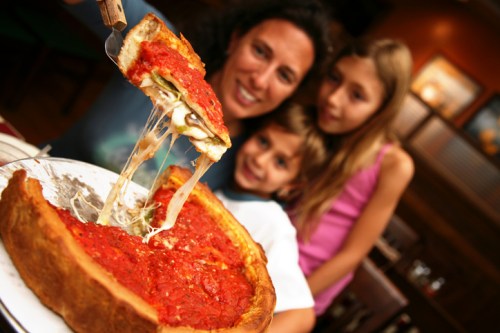 A woman and two children enjoy a cheesy deep-dish pizza, with gooey cheese stretching from a slice.