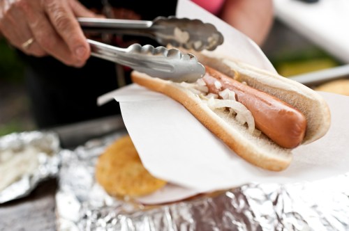 A person using tongs to place a hot dog with onions into a bun, resting on a piece of white paper.