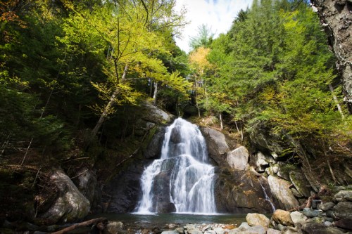A serene waterfall cascades down rocky cliffs, surrounded by lush green trees and autumn foliage.