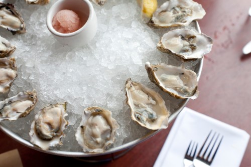A platter of fresh oysters on ice, served with a small cup of sauce and a lemon wedge.