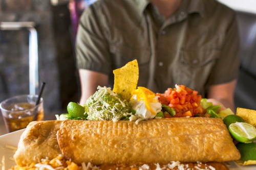 A large plate of enchiladas topped with guacamole, pico de gallo, and a side of chips, with a drink in the background.