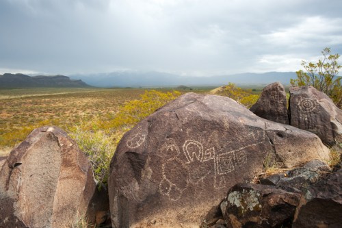 Petroglyphs etched on large rocks, set against a vast desert landscape under a cloudy sky.
