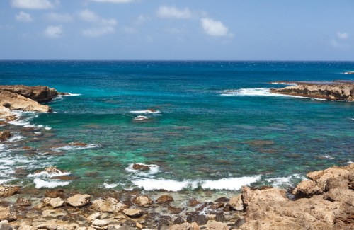 A serene coastal view featuring clear turquoise waters, rocky shores, and a bright blue sky with scattered clouds.