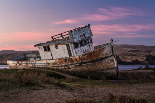 An old, weathered boat rests on land at sunset, surrounded by grass and hills under a colorful sky.