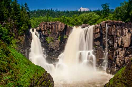 A scenic waterfall cascading over rocky cliffs, surrounded by lush greenery and a clear blue sky.