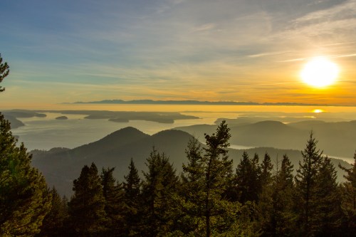 A scenic view of mountains and water at sunset, with trees in the foreground and a colorful sky.