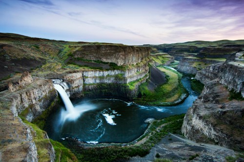 A stunning waterfall cascades into a winding river, surrounded by rugged cliffs and lush greenery under a twilight sky.