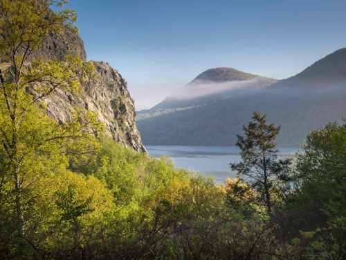 A serene landscape featuring lush green trees, rocky cliffs, and a misty river valley under a clear blue sky.