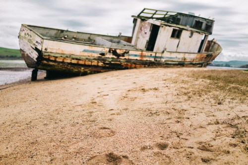 An abandoned, weathered boat rests on a sandy shore, partially submerged, with footprints in the foreground.