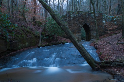 A serene forest scene featuring a stone arch bridge over a flowing stream, surrounded by trees and autumn foliage.