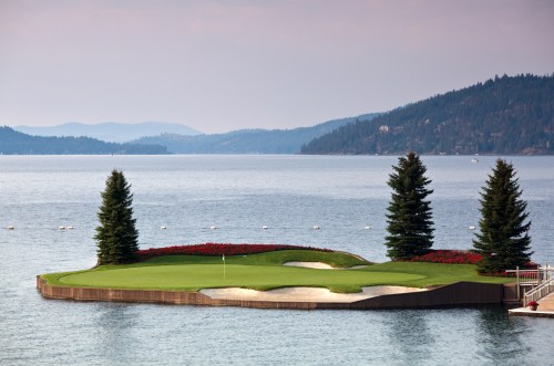 A scenic golf course island surrounded by water, featuring sand traps and lush greenery against a mountainous backdrop.