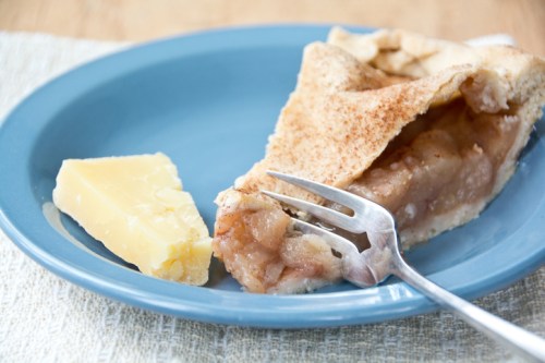 A slice of apple pie on a blue plate, accompanied by a piece of yellow cheese and a fork.