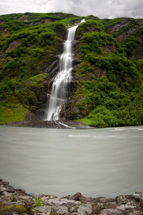 A cascading waterfall flows down a rocky hillside into a calm, gray river surrounded by lush greenery.