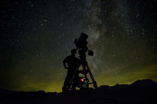 A silhouette of a person observing the night sky with a telescope, surrounded by stars and the Milky Way.