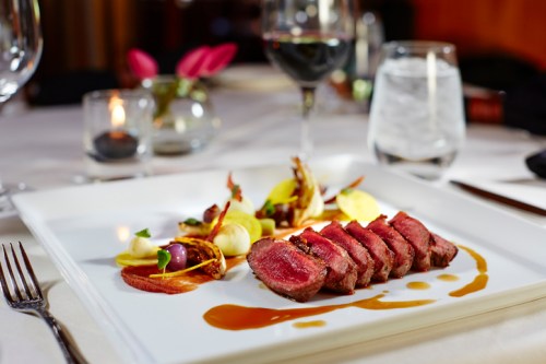 A gourmet plate featuring sliced steak, colorful vegetables, and a drizzle of sauce, accompanied by wine and water glasses.