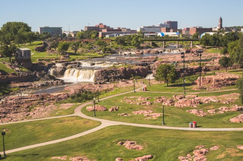 A scenic view of a park with waterfalls, rocky terrain, and city buildings in the background under a clear blue sky.