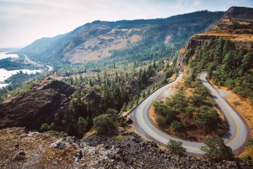 A winding road curves through a lush green landscape, surrounded by mountains and a river in the distance.