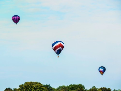 Three colorful hot air balloons float in a clear sky above green trees.