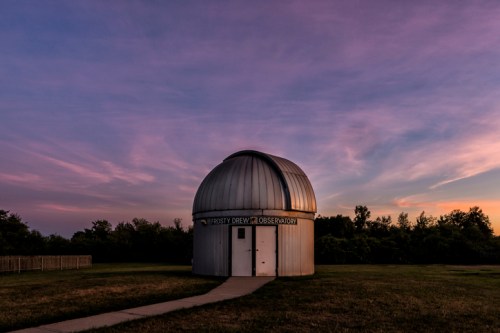 A silver observatory dome at dusk, surrounded by grass and trees under a colorful sky.