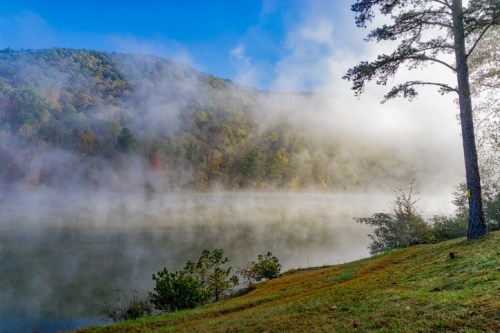 A serene lake surrounded by misty hills and trees, with a clear blue sky above.