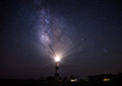 A lighthouse beams light under a starry sky, with the Milky Way visible in the background.
