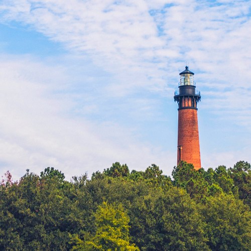 A tall lighthouse stands above lush green trees under a partly cloudy sky.