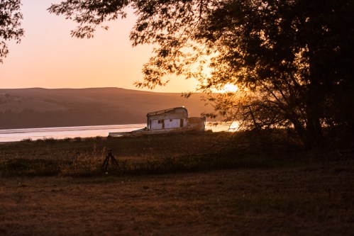 A weathered boat rests on the shore at sunset, surrounded by trees and reflecting golden light on the water.