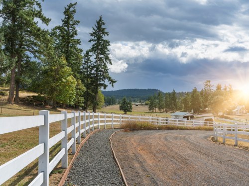 A winding gravel path lined with a white fence, surrounded by trees and fields under a cloudy sky with a sunset glow.