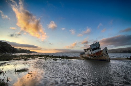 An abandoned boat rests on a muddy shore at sunset, with clouds reflecting in the water and hills in the background.
