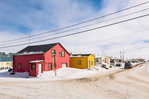 A snowy street scene featuring a pink house and a yellow house, with power lines and vehicles in the background.