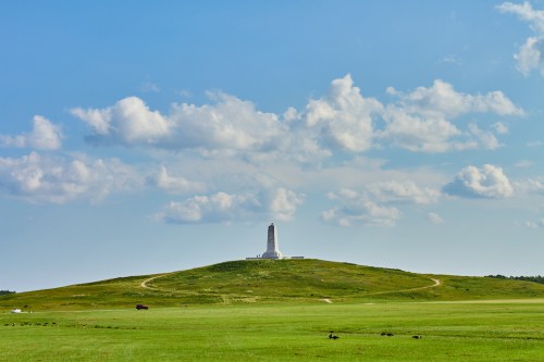 A tall monument on a grassy hill under a blue sky with scattered clouds. A winding path leads to the monument.
