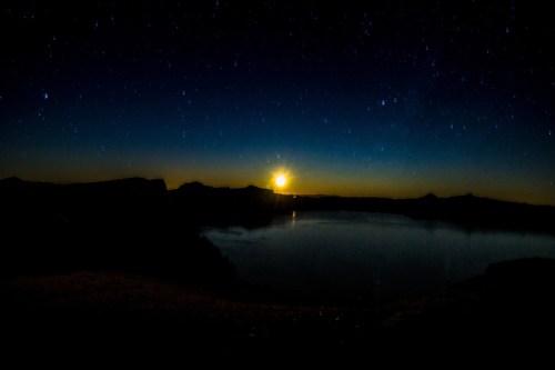A serene night sky with stars above a calm lake, illuminated by a rising sun on the horizon.