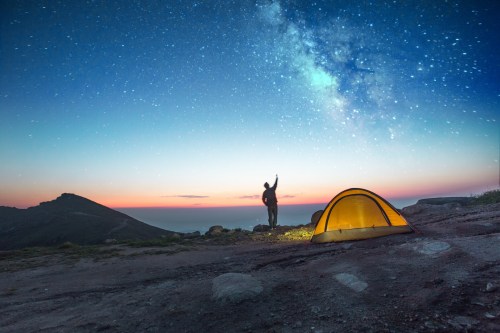 A camper stands by a glowing tent under a starry sky, with mountains and ocean in the background at dusk.